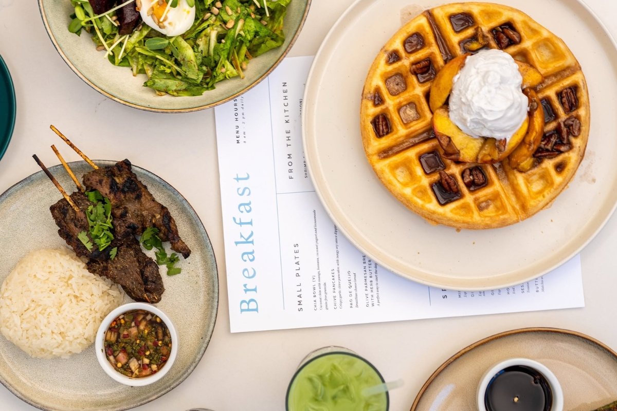 Overhead flatlay of Gati Cafe breakfast menu including a pecan waffle with whipped cream, grilled meat skewers with rice, a green salad with soft-boiled egg, and a green smoothie on East Holly St in East Austin